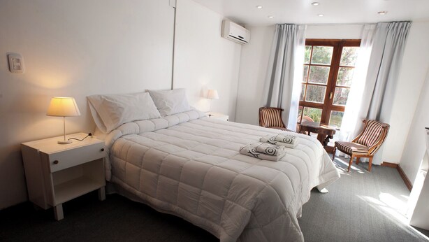 A neatly made bed with a white duvet, a bedside table with a lamp, a chair, and a window with curtains. - Casa Bonita (Neuquén)