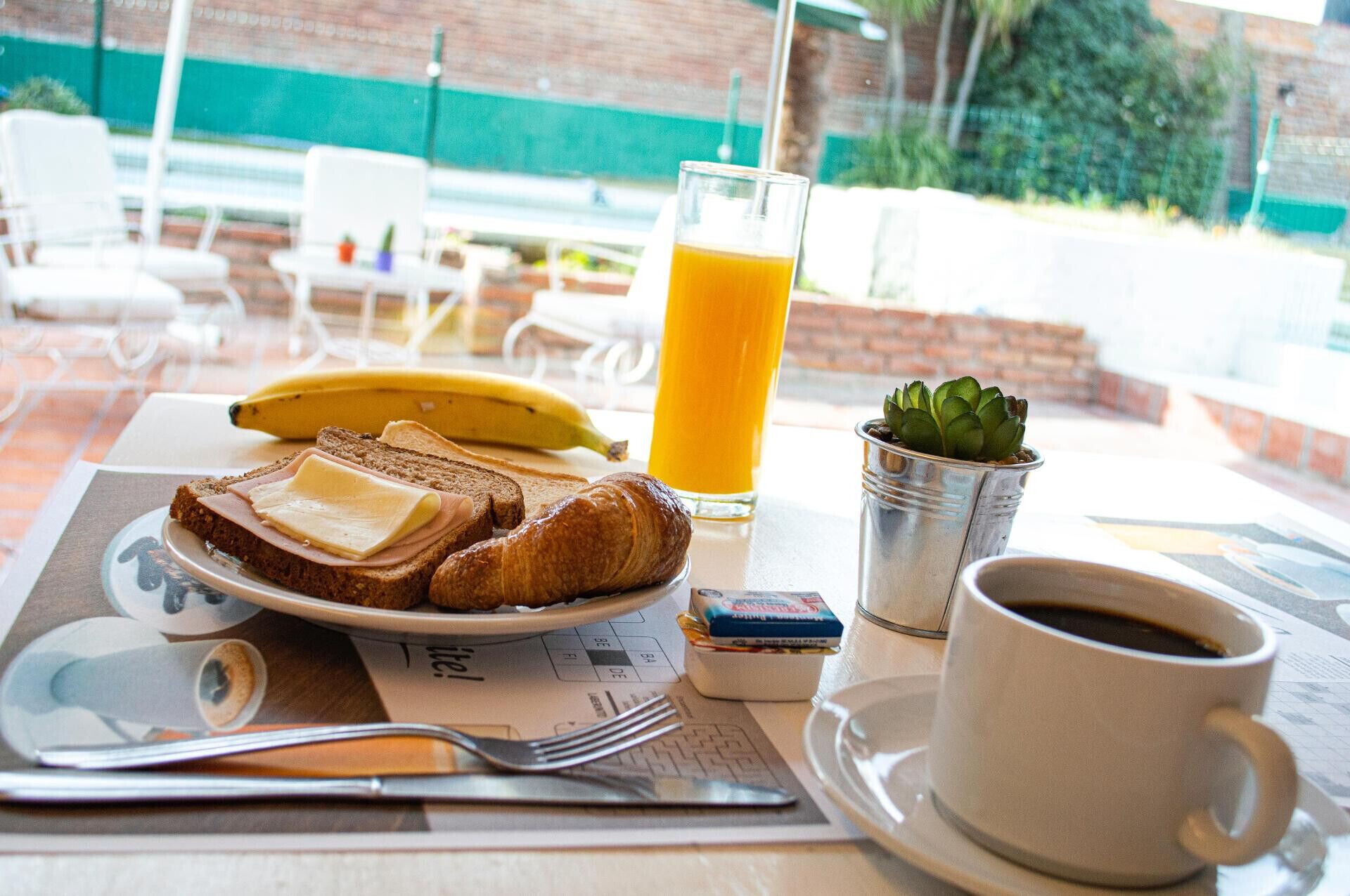 Una escena de desayuno con tostadas, un plátano, un vaso de jugo de naranja, una taza de café y una planta pequeña sobre una mesa.