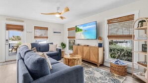 A living room with a blue sofa, a wooden cabinet, a television, and a large window with a view of the beach. - 197 Beach Daze: 3BR Pool Home w/ Deeded Beach Access! (Fort Myers Beach)