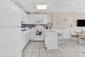 A modern kitchen with white cabinetry, a built-in microwave, and a dining area with white chairs and a wooden table. - Seagull Heights! 2-BR 2-BA OCEANFRONT Royal Garden Resort Condo In&Outdoor Pools (Murrells Inlet)