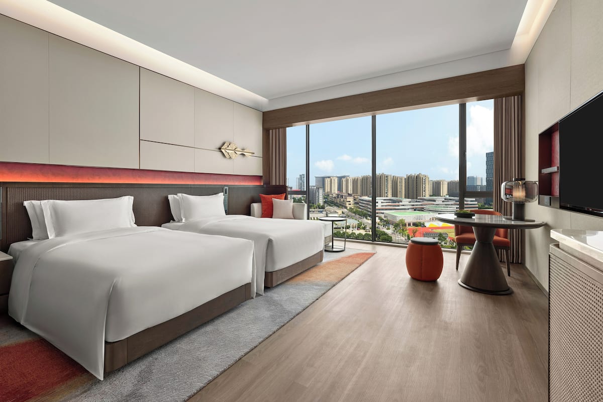 A modern hotel room with two beds, a dining area, and a large window offering a city view.