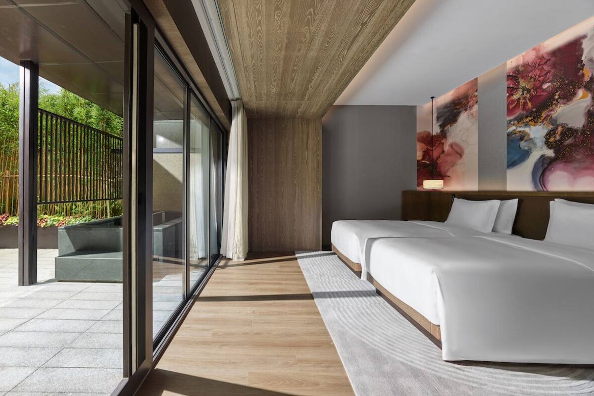 A modern hotel room with a large bed, a seating area, and a view of greenery outside.