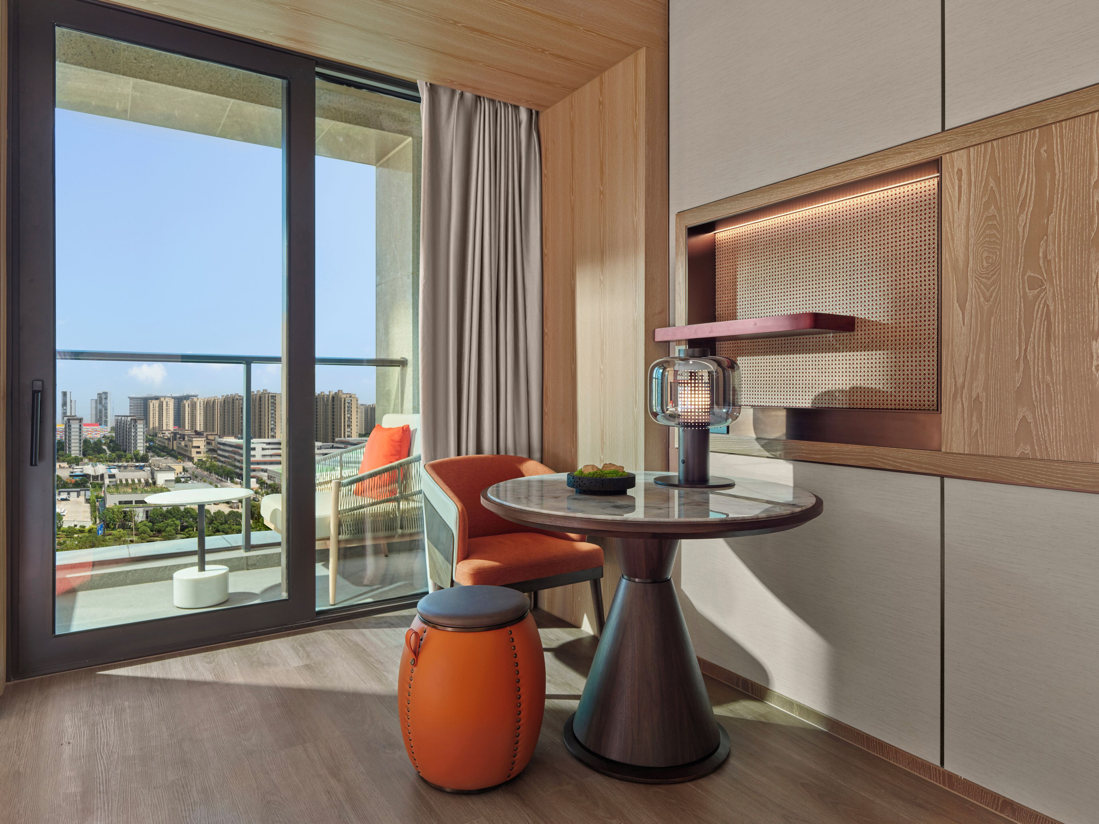 A modern hotel room with a large balcony, a round table, a chair, and a small orange stool.