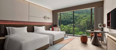Een moderne hotelkamer met twee bedden, een groot raam met uitzicht op een bos, een zithoek en een televisie.