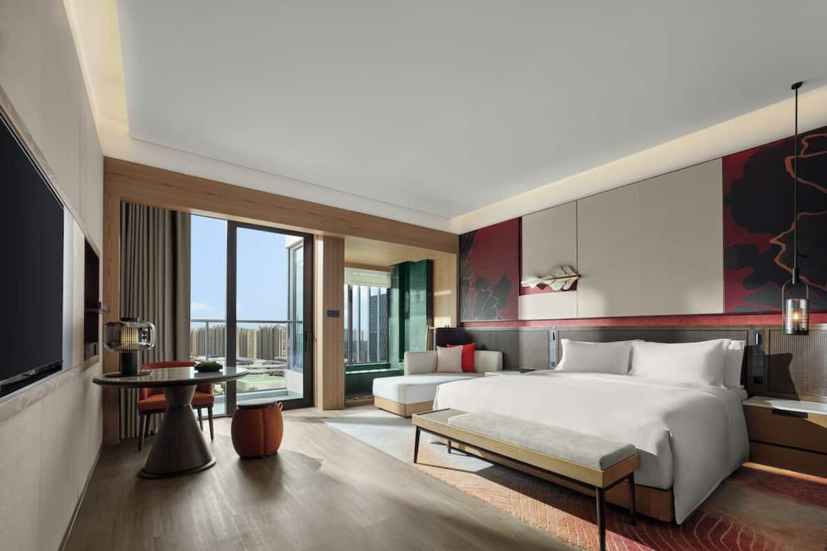 A modern hotel room with a large bed, a desk with a chair, a dining table, and a view of the city.