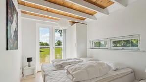 Une chambre à coucher comprenant un lit, une table de chevet, une lampe, une fenêtre avec des stores et une vue sur la verdure à l’extérieur.