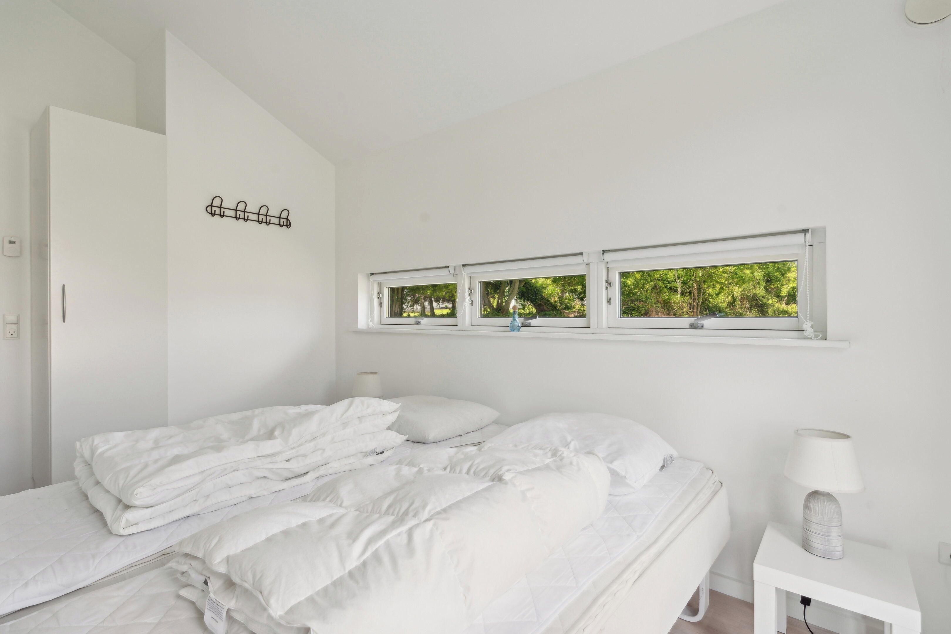 Una camera da letto bianca con un letto, un comodino con una lampada, una finestra vista sul verde e un appendiabiti.