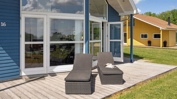 Une maison moderne dotée de grandes portes vitrées, d’une terrasse en bois et de deux fauteuils de relaxation.