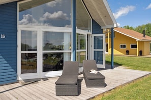 Une maison moderne dotée de grandes portes vitrées, d’une terrasse en bois et de deux fauteuils de relaxation.