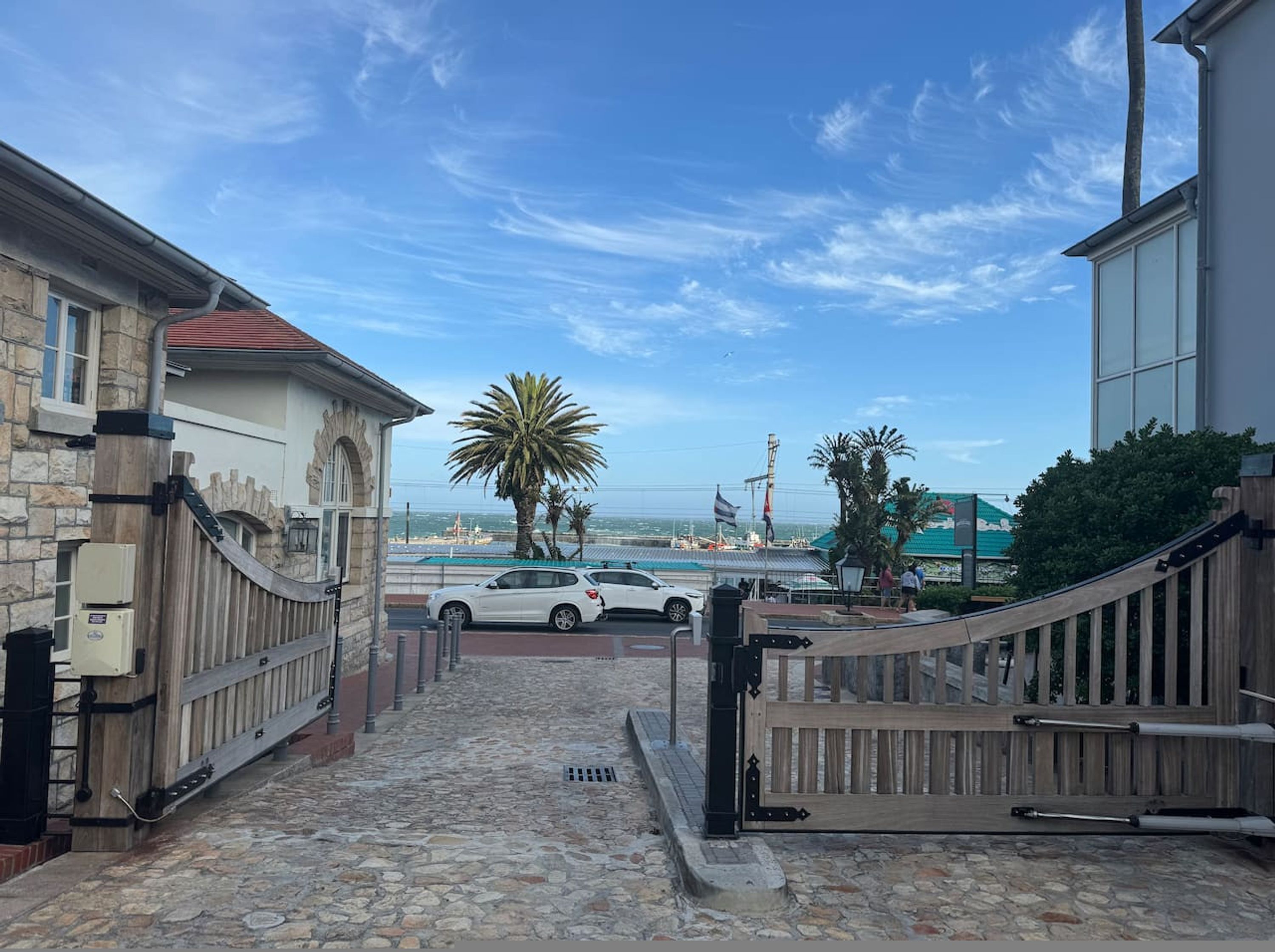 A stone-paved pathway leading to a gated entrance with a view of the sea and boats in the background.