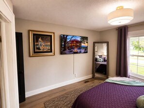 Un dormitorio con cama, televisor montado en la pared, cuadro enmarco, espejo y ventana con cortinas.