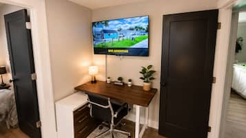 Habitación de hotel con escritorio, televisor montado en la pared, una planta pequeña en maceta y una cama con cubrecama blanco.