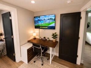 A hotel room with a desk, a television mounted on the wall, a small potted plant, and a bed with a white bedspread.