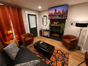 A modern living room with a fireplace, two leather armchairs, a black sofa, and a cityscape TV.