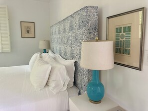 Een net opgemaakt bed met witte lakens, een hoofdbord met een patroon, een blauwgroene lamp en een lijst met kunstwerk aan de muur.