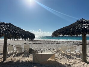 Een strand met twee rieten parasols, een tafel met tafelkleed en meerdere witte ligstoelen.
