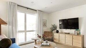A modern living room with a sofa, coffee table, TV, and a large window with curtains. - Tide House  | Manhattan Beach - Unit A - Seaside w/ Parking (Manhattan Beach)