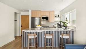 A modern kitchen with a central island, wooden bar stools, stainless steel appliances, and a view of the adjacent living area. - Tide House  | Manhattan Beach - Unit A - Seaside w/ Parking (Manhattan Beach)