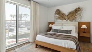 A bedroom with a large bed, two bedside lamps, a window with curtains, and a view of palm trees and a parking lot. - Tide House | Manhattan Beach - Unit B - Seaside w/ Parking (Manhattan Beach)