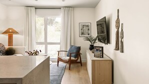 A modern living room with a large window, a sofa, a chair, a coffee table, and a TV on the wall. - Tide House | Manhattan Beach - Unit C - Seaside w/ Parking (Manhattan Beach)