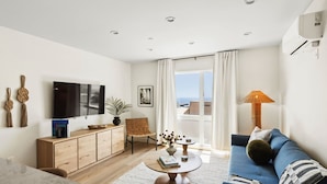 Una sala moderna con televisor de pantalla plana, muebles de madera, un sofá azul, una mesa de centro redonda y vista al mar a través de grandes ventanales.