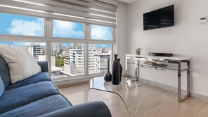 A modern living room with a sofa, a glass coffee table, a TV mounted on the wall, and a view of the city through large windows.