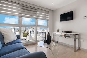 Una sala de estar moderna con un sofá, una mesa de centro de vidrio, un televisor montado en la pared y vistas a la ciudad a través de amplios ventanales.
