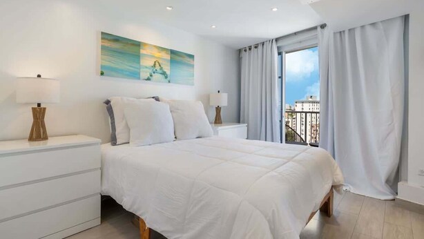 A modern bedroom with a large bed, two bedside tables with lamps, a painting on the wall, and a balcony with curtains.