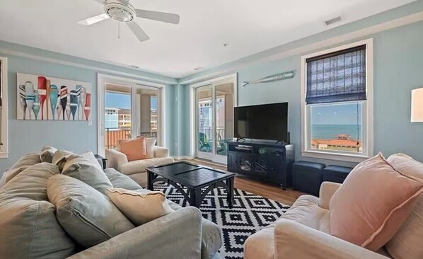 A living room with a sofa, coffee table, and TV. There are doors leading to a balcony with a view of the ocean.