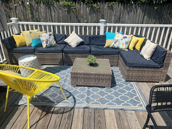 Un canapé d’angle en osier agrémenté de plusieurs coussins, une petite table ronde avec une plante, une chaise jaune et une chaise noire, le tout placé sur une terrasse en bois dotée d’une rambarde blanche.
