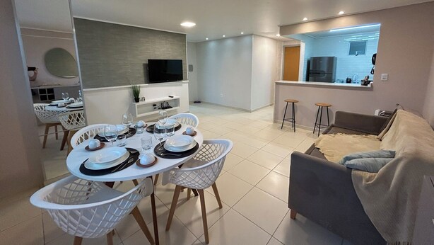 A modern living room with a grey sofa, a dining table set for four, and a kitchen area with a TV and bar stools.