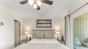 A bedroom with a bed, two bedside tables with lamps, a ceiling fan, a painting, and a sliding glass door leading to a balcony.