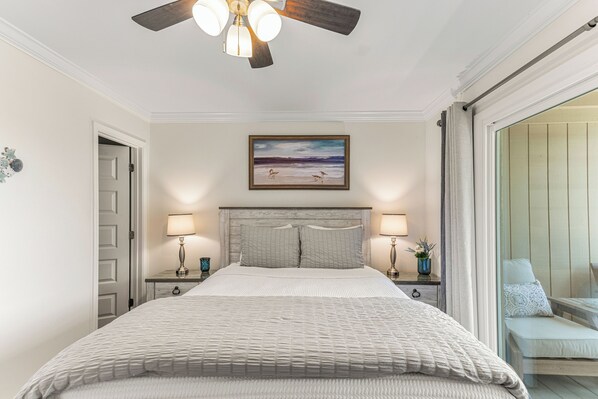 A bedroom with a bed, two bedside tables with lamps, a ceiling fan, a painting, and a sliding glass door leading to a balcony. - Pristine Oceanview Escape + Community Pool (Tybee Island)
