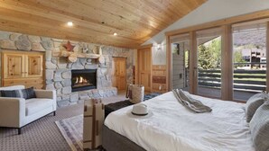 Camera da letto con camino in pietra, soffitto in legno, un letto, un divano e vista panoramica attraverso ampie vetrate.