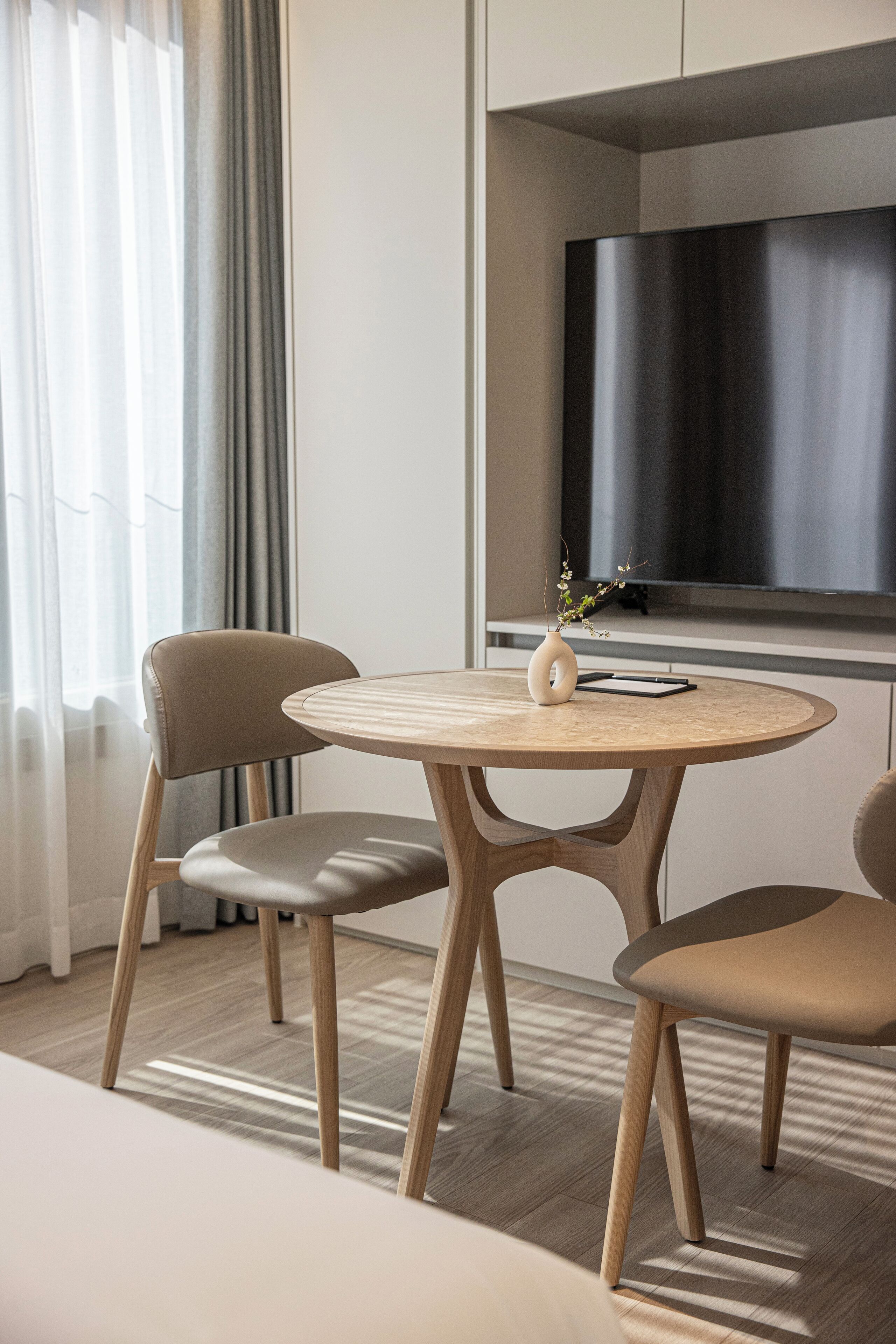 Habitación de hotel moderna con una mesa redonda de madera, dos sillas, un florero con flores, un televisor de pantalla plana y un ventanal grande con cortinas.