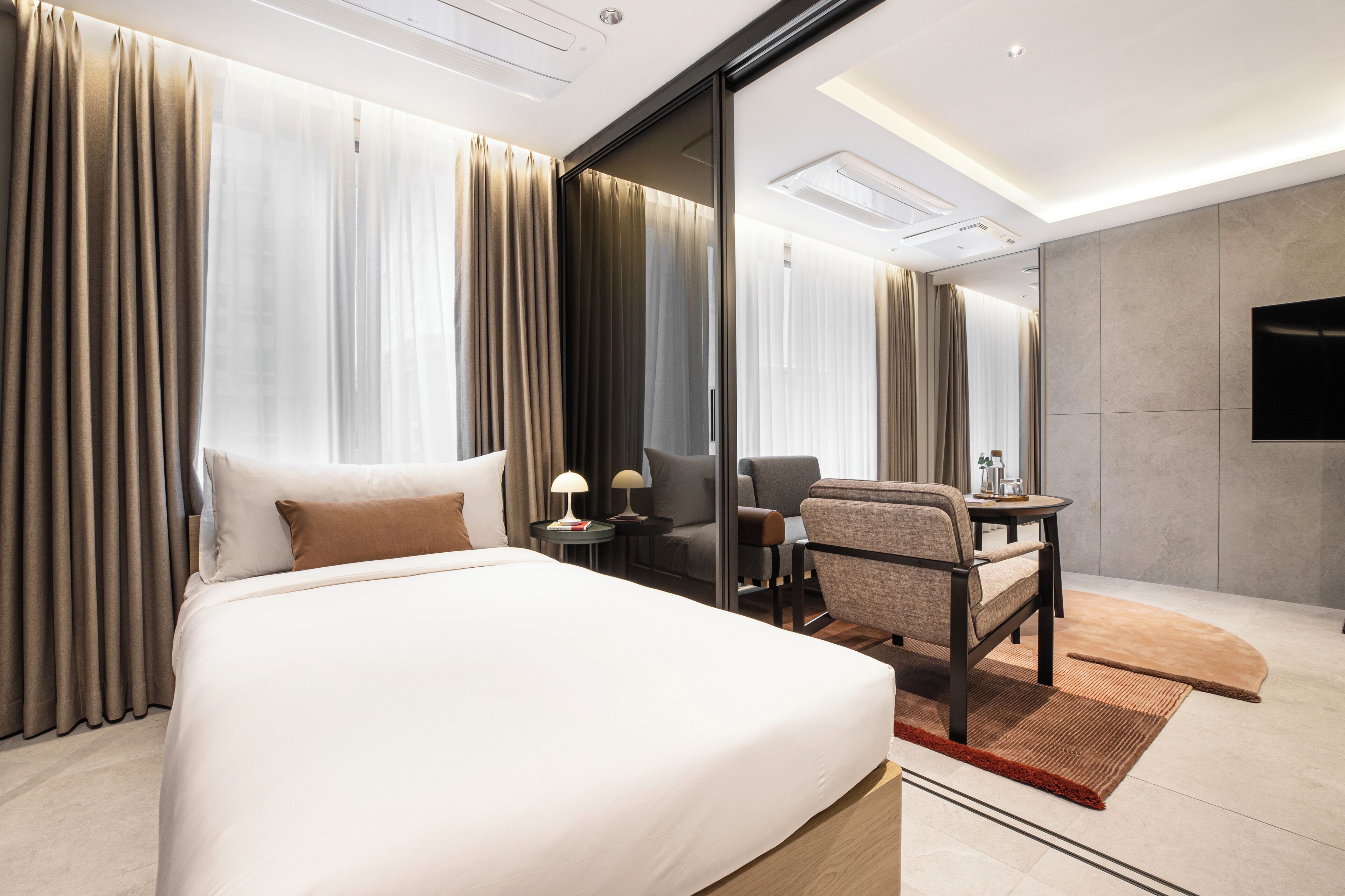 A modern hotel room with a large bed, a seating area with a chair and table, and a flat-screen TV mounted on the wall.