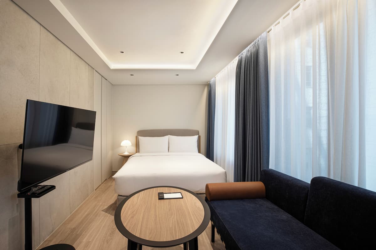 A modern hotel room with a flat-screen TV, a bed, a sofa, a round table, and a large window with curtains.