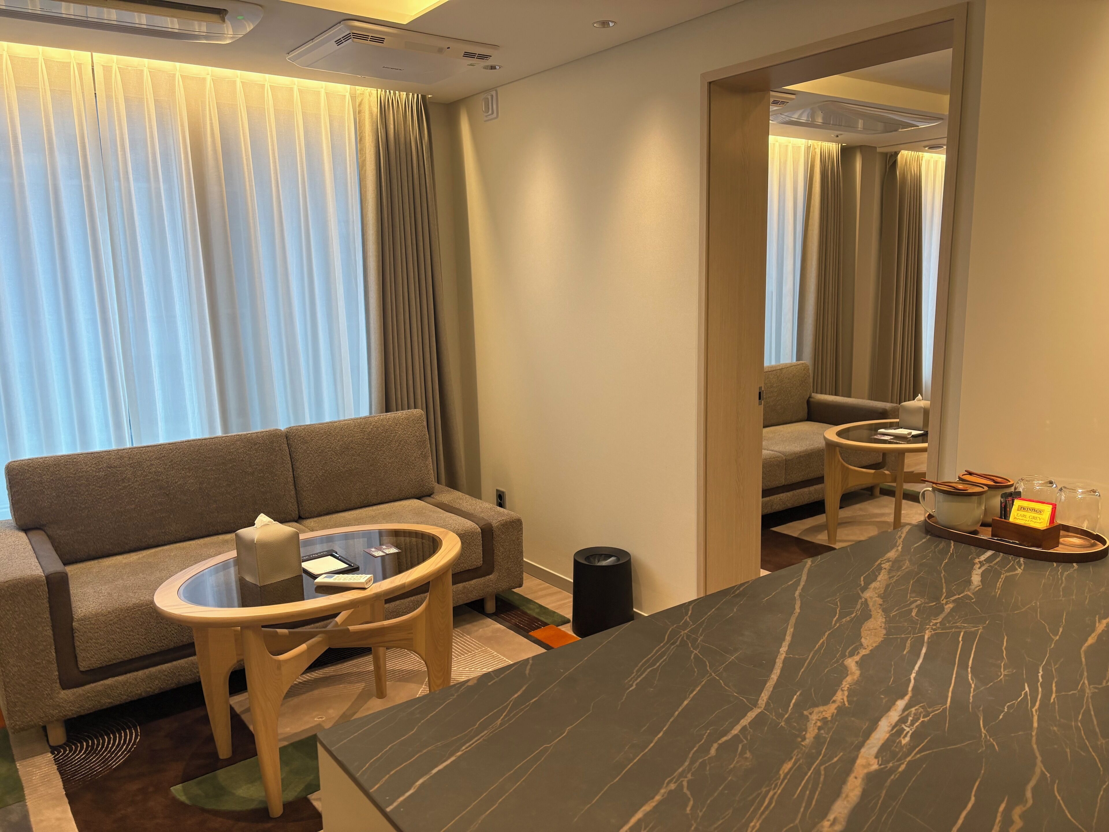 Habitación de hotel con sofá, una pequeña mesa redonda, una mesita de centro, un espejo y una bandeja con cafetera y un cartel.