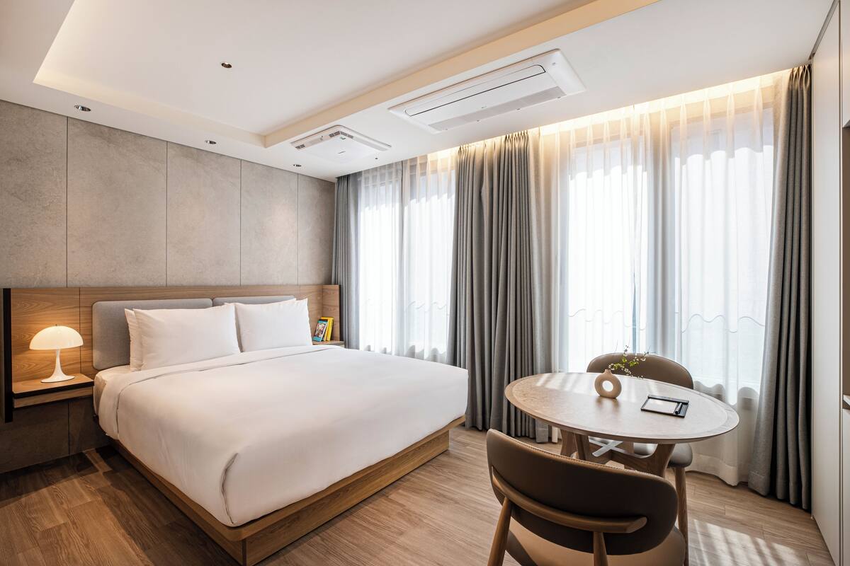 A modern hotel room with a large bed, bedside table, lamp, and a round table with chairs.