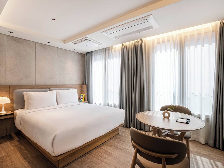 A modern hotel room with a large bed, bedside table, lamp, and a round table with chairs.