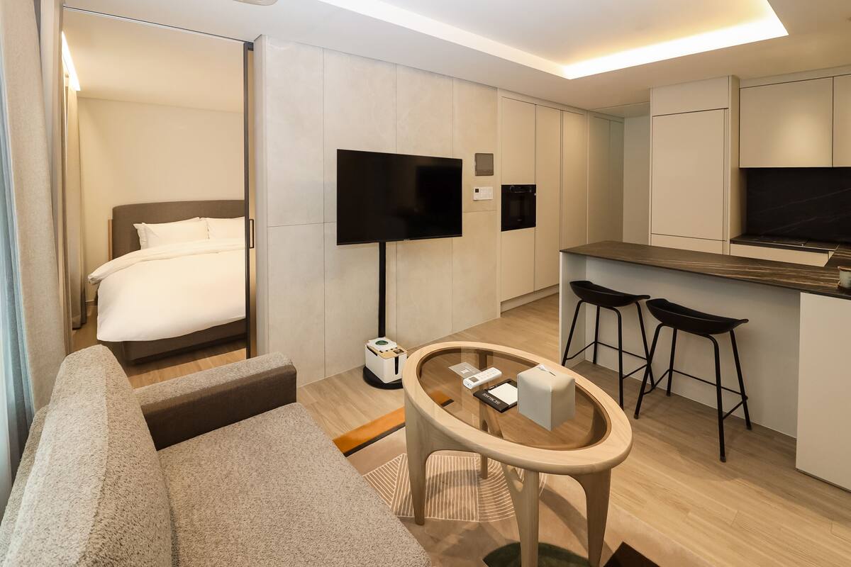 A modern hotel room with a bed, a TV, a small kitchen area, and a seating area.