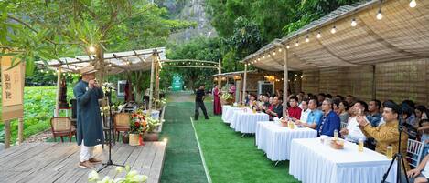 An outdoor event with a stage, audience, and tables set up under a covered area with string lights.