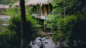 Une terrasse en bois surmontée d’une structure au toit de chaume, entourée d’une végétation luxuriante et bordée par un plan d’eau.