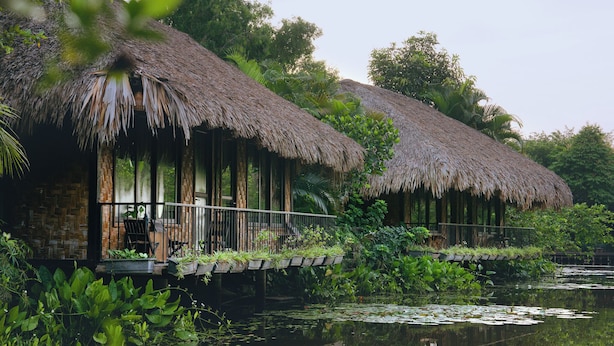 Un bungalow au toit de chaume, doté d’une terrasse en bois surplombant l’eau, et entouré d’une végétation luxuriante.