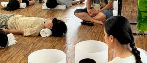 A room with wooden flooring, a large window, and a view of greenery outside. There are several people lying on the floor, some sitting, and a person holding a bowl.
