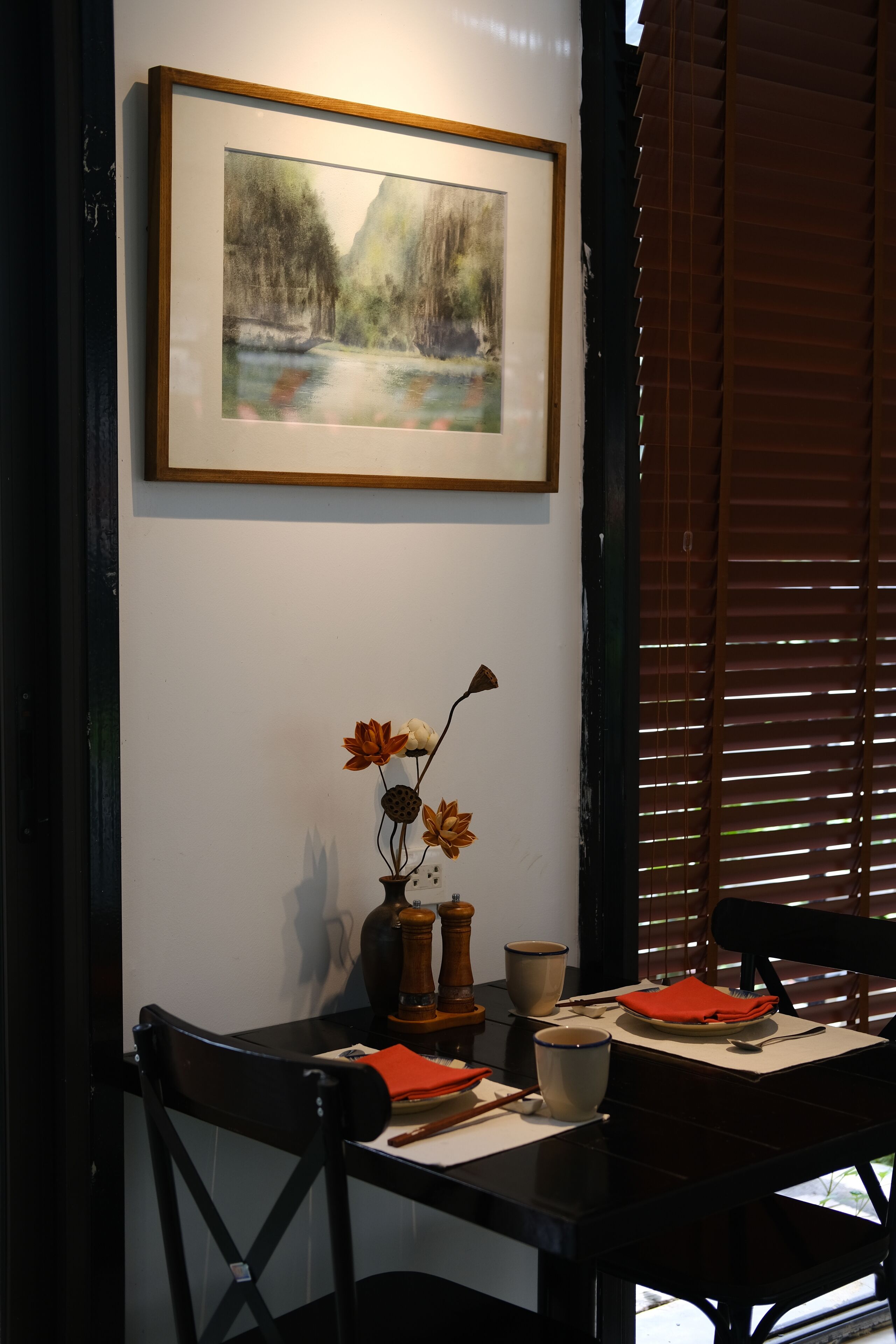 A dining area with a table set for two, a framed painting on the wall, and a vase with flowers on the table.