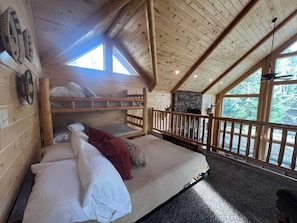 Cozy cabin-style bedroom with a bunk bed arrangement, stone fireplace, and large windows offering forest views.