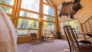 L’intérieur d’une cabane en rondins, avec de grandes fenêtres, un tapis en fourrure sur le sol et une peau d’animal empaillée accrochée au mur.