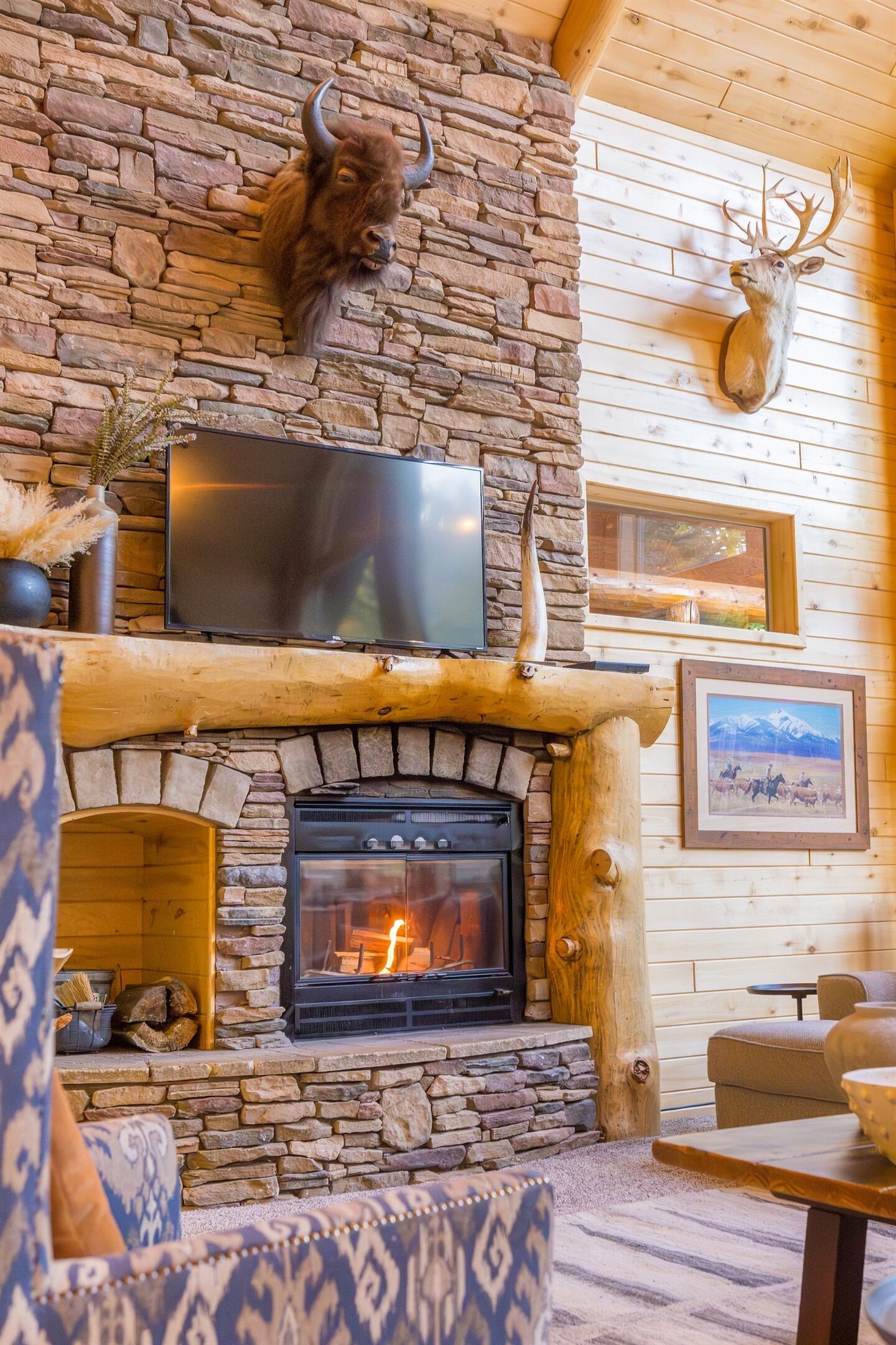 A stone fireplace with a TV above it, mounted on a wooden wall with a stone bull head and a mounted deer head.