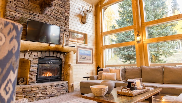 L’intérieur chaleureux d’un chalet avec une cheminée en pierre, une tête de cerf accrochée au mur, un téléviseur et une grande fenêtre donnant sur la forêt.
